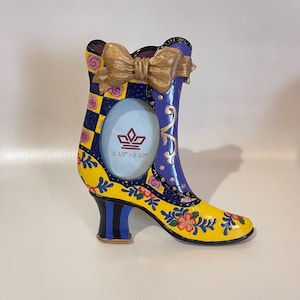 May include: A decorative boot-shaped picture frame with a bow on top. The frame is yellow, blue, and purple with floral and checkered patterns. The photo opening is 2 1/2" x 3 1/2".