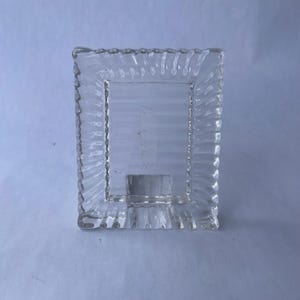 May include: Clear glass picture frame with a rectangular opening and a textured, wavy border. The frame has a small, square base and is designed to stand upright. The glass is transparent, allowing light to pass through.