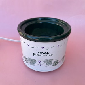May include: A white Rival Potpourri Crock electric warmer with a dark green interior rim. The exterior features a painted vine and berry pattern around the base and top. A white power cord extends from the side of the cylindrical ceramic vessel.