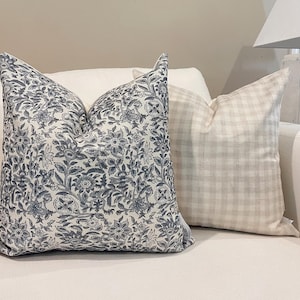 Könnte beinhalten: Zwei dekorative Kissen auf einem weißen Sofa. Ein Kissen hat ein blau-weißes Blumenmuster, das andere ein neutrales Karomuster. Diese Kissen verleihen dem Wohnraum einen Hauch von Eleganz.