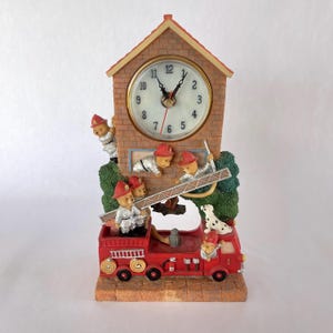 Puede incluir: Un reloj decorativo que representa un camión de bomberos rojo con osos de peluche bomberos. El reloj tiene una fachada de estilo ladrillo con una esfera y un pequeño techo en forma de casa. También hay un perro dálmata.