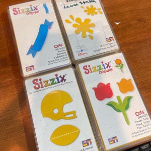 May include: Four Sizzix Originals die-cut sets. The sets include a blue banner, a yellow splatter, a yellow American football helmet and ball, and a red and orange tulip with green leaves. Each set is in a clear plastic case.