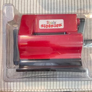 May include: A red Sizzix Sidekick die-cutting and embossing machine in its original clear plastic packaging. The machine has a black handle and base. The Sizzix Sidekick logo is visible on a white label.