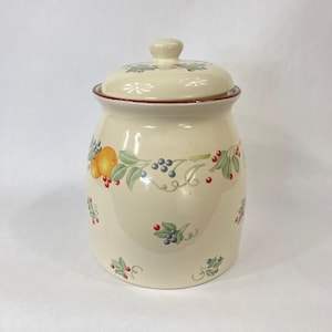 Op de afbeelding: Crèmekleurige keramische pot met deksel, versierd met een fruit- en bessenontwerp. De pot heeft een rode rand en een kleine knop op het deksel. Het fruitontwerp omvat sinaasappels, blauwe bessen en rode bessen met groene bladeren.