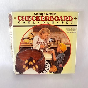 May include: A vintage "Chicago Metallic CHECKERBOARD CAKE PAN SET" box. The box features a picture of a checkerboard cake and a woman and child in a kitchen. The box is cream-colored with red and brown text and graphics.