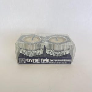 May include: A clear plastic package containing two crystal tea light candle holders. Each holder contains a small, round candle. The package has the text "Brite Lite Crystal Twin Tea Light Candle Holders".