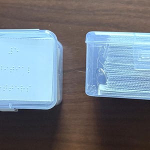 Könnte beinhalten: Zwei transparente Kunststoffbehälter auf einer braunen Oberfläche. Ein Behälter enthält einen Stapel weißer Karten mit Blindenschrift und einen Schlüsselanhänger. Der andere Behälter ist mit einem Stapel dünner, silberfarbener Drähte gefüllt.