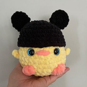 May include: A crocheted yellow chick toy with pink feet and black ears. The chick is wearing a black crocheted hat with two black pom-poms on top.