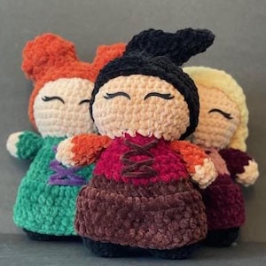 May include: Three crocheted plush dolls, each with a different colored dress and hair. The doll on the left is wearing a green dress and has red hair. The doll in the middle is wearing a burgundy dress and has black hair. The doll on the right is wearing a burgundy dress and has blonde hair.