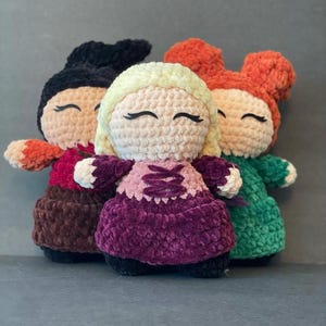 Pode incluir: Três bonecas de pelúcia de crochê com cabelo e roupas de cores diferentes. A boneca à esquerda tem cabelo preto e uma roupa vermelha e marrom. A boneca do meio tem cabelo loiro e um vestido roxo. A boneca à direita tem cabelo laranja e um vestido verde.