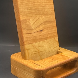 May include: A wooden phone stand with a light brown finish. The stand has a rectangular base with rounded edges and a slot to hold a phone. The back piece is angled for optimal viewing.
