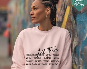 Op de afbeelding: Een lichtroze sweatshirt met de tekst "Let them" en een inspirerende boodschap in zwart schrift. De sweatshirt wordt gedragen door een persoon met gevlochten haar en gouden oorringen, leunend tegen een met graffiti bedekte muur.