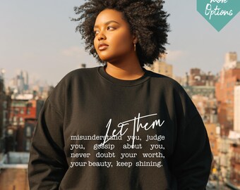 Op de afbeelding: Zwarte sweatshirt met de tekst "Let them" in een sierlijk lettertype. Daaronder staat de tekst: "misunderstand you, judge you, gossip about you, never doubt your worth, your beauty, keep shining."