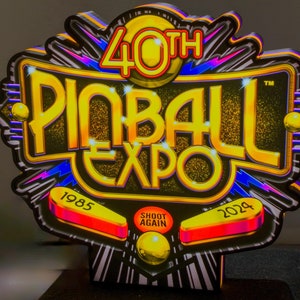 May include: A colorful sign for the 40th Pinball Expo, featuring a gold and black design with the text "Pinball Expo" in large, stylized letters. The sign includes the years 1985 and 2024, and the words "Shoot Again".