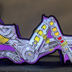 Può includere: Oggetto decorativo a tema robot, con una combinazione di colori viola e argento. Il design presenta una figura robotica stilizzata con dettagli meccanici gialli e rossi. L'oggetto è posto su uno sfondo scuro.
