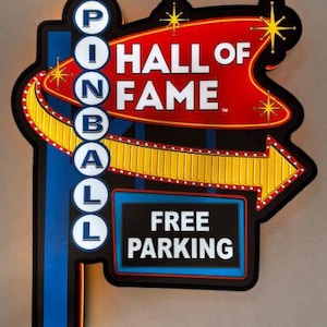 Könnte beinhalten: Retro-Schild für eine Pinball Hall of Fame mit den Worten "PINBALL HALL OF FAME" in weißer Schrift auf einem roten und gelben Pfeildesign. Das Schild enthält auch "FREE PARKING".