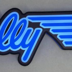 以下が含まれることがあります： 黒と青のバリーの看板。キーの形をしており、「Bally」の文字は青色の筆記体で、青色の縁取りがされています。キーのデザインは青色のストライプと黒色の円で構成されています。