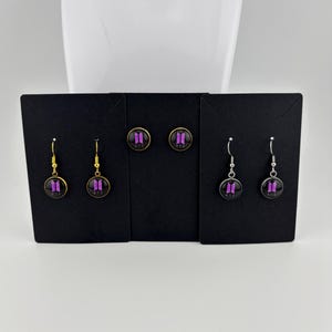 Puede incluir: Tres pares de pendientes circulares presentados sobre tarjetas negras. Un par tiene ganchos dorados, un par consiste en pendientes de botón con montura dorada y un par tiene ganchos plateados. Cada pendiente muestra un logo BTS morado.