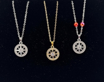 May include: Three necklaces with compass pendant designs. One is silver with a clear stone, another is gold with a clear stone, and the third is silver with a clear stone and two red beads.
