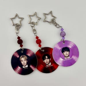 May include: Three circular keychains featuring portraits of Jimin, Taehyung, and Jung Kook on vinyl record-style discs. Each keychain has a silver star-shaped clasp, a metal link chain, and decorative beads in red or purple colors.