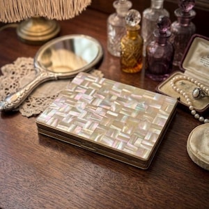 May include: A vintage vanity scene featuring a mother-of-pearl compact, a hand mirror, and several antique perfume bottles. The compact has a rectangular shape with a gold-colored frame. A pearl necklace and ring are visible in a small jewelry box.