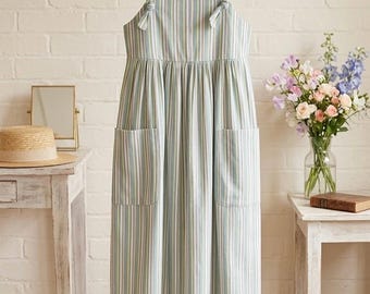 May include: A sleeveless midi dress featuring a vertical striped pattern in shades of sage green, white, yellow, and lavender. The dress has a square neckline with small decorative knots at the straps and two large patch pockets on the front skirt.