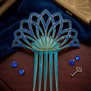 May include: A hair comb featuring a fan-shaped top with a teal enamel finish and intricate cutouts. The design is accented with small, embedded blue crystals along the edges. The comb has five long, slender prongs extending from the base.