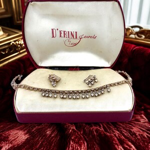 May include: An open burgundy jewelry box with a cream-colored interior displays a necklace and earrings set. The jewelry is silver-toned with clear gemstones. The box lid has the text "D'ERINI Jewels by fiori" in red script.