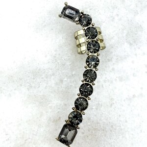 May include: A gold-toned ear cuff featuring a curved row of dark gray, faceted circular and rectangular crystals. The design includes a multi-banded section that wraps around the ear cartilage, accented with a series of prong-set stones.