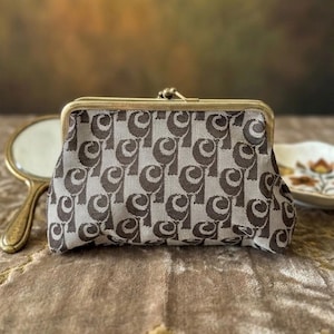 May include: A small, vintage coin purse with a gold-toned metal frame and clasp. The purse features a repeating pattern of brown and white abstract shapes. A small, gold-toned hand mirror and a decorative plate are visible in the background.