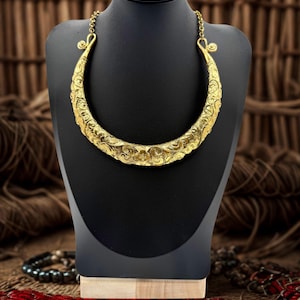 May include: A gold-colored statement necklace with an ornate, engraved design. The necklace is displayed on a black mannequin, showcasing its curved shape and intricate details. The background features a rustic wooden texture.