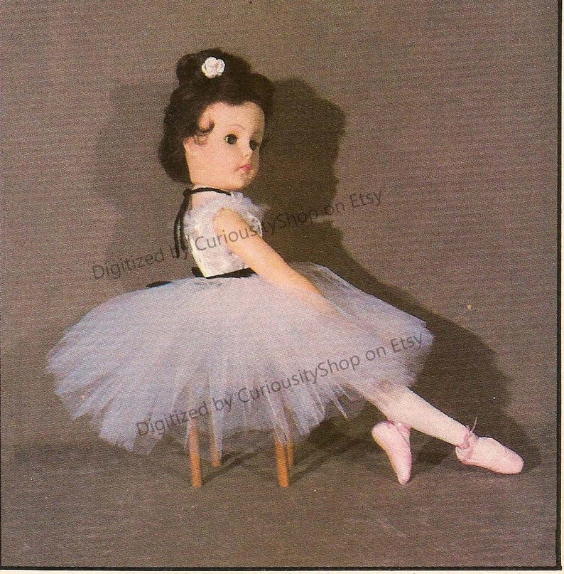 May include: A porcelain doll dressed in a white tulle tutu and a black and white bodice. The doll is sitting on a wooden stool with one leg extended and her foot pointed. The doll has brown hair and a white flower in her hair.