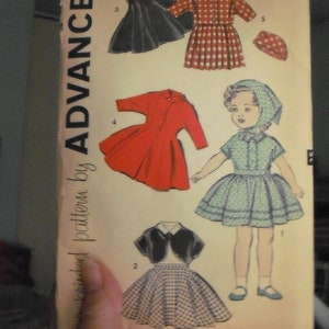 May include: A vintage sewing pattern for a 12-inch doll. The pattern includes five different outfits: a black dress with a white collar, a red dress, a plaid dress, a green dress, and a black jacket with a white collar and a plaid skirt. The pattern is titled "9211 Doll Size 12 Inch" and is from "Advance Pattern by Advance".