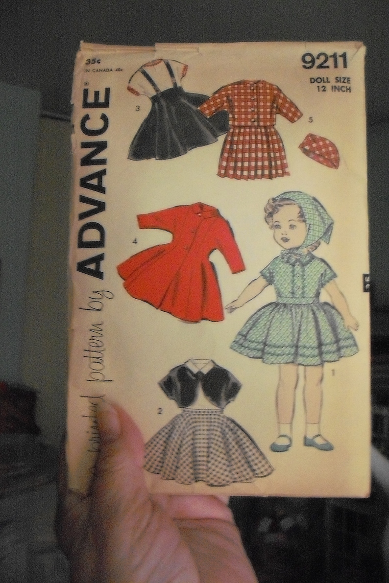 May include: A vintage sewing pattern for a 12-inch doll. The pattern includes five different outfits: a black dress with a white collar, a red dress, a plaid dress, a green dress, and a black jacket with a white collar and a plaid skirt. The pattern is titled "9211 Doll Size 12 Inch" and is from "Advance Pattern by Advance".