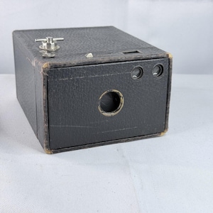 May include: A vintage black box camera with a textured leatherette finish. The front features a central circular lens opening and two smaller circular viewfinders. A silver metal winding key is positioned on the top surface of the camera body.