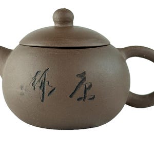 May include: A brown ceramic teapot with a rounded body, a curved handle, and a small spout. The lid has a small knob. The teapot has black Chinese characters on the side. The teapot is designed for brewing tea.