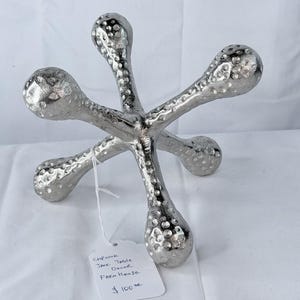 May include: A silver-colored, decorative jack table decor with a textured surface. The object has six rounded arms extending from a central point. A tag attached to the object reads "Chrome Jack Table Decor Farm House".
