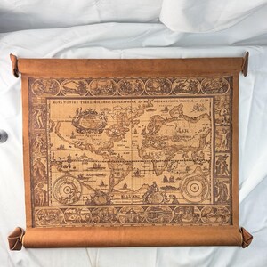 May include: A brown leather scroll featuring a vintage-style world map titled Nova Totius Terrarum Orbis Geographica Ac Hydrographica Tabula. The map is surrounded by decorative illustrations of figures and scenes in a rectangular border.