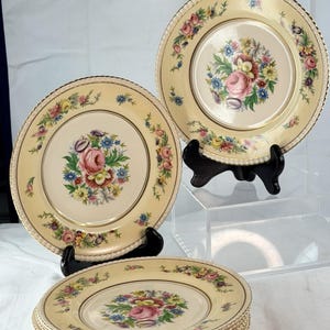 May include: A collection of vintage floral plates. The plates have a cream-colored background with a central floral bouquet design in pink, blue, yellow, and green. The rims are adorned with a delicate gold trim and a repeating pattern. Some plates are displayed on black stands.