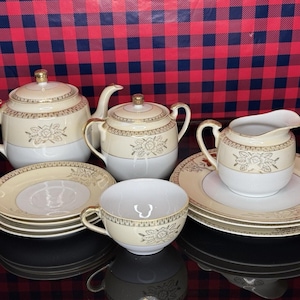 May include: A vintage tea set with a cream and white color scheme, featuring gold accents and floral designs. The set includes a teapot, sugar bowl, creamer, teacups, and saucers. The tea set is displayed on a reflective surface.