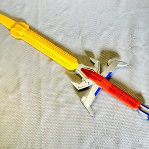 Può includere: Una spada giocattolo gialla e rossa con dettagli argentati. La spada presenta un'impugnatura e una lama gialle, una sezione centrale rossa e una guardia argentata. Il giocattolo ha un design futuristico.