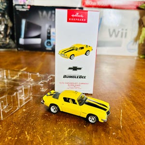 Könnte beinhalten: Ein gelbes 1976 Chevrolet Camaro Bumblebee Druckgussmodellauto mit schwarzen Rennstreifen. Das Auto wird neben seiner Hallmark Keepsake-Box präsentiert. Die Box zeigt ein Bild des Autos und das Chevrolet-Logo.