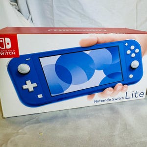May include: A rectangular cardboard box for a blue Nintendo Switch Lite handheld gaming console. The box features the Nintendo Switch logo in the top left corner and the text Nintendo Switch Lite in the bottom right corner, with a graphic.