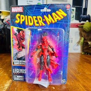 K&ouml;nnte beinhalten: Eine Marvel Spider-Man Legends Series Actionfigur von Elektra Natchios, Daredevil, in einer transparenten Kunststoffverpackung. Die Figur ist rot und schwarz, mit zwei silbernen Sai-Waffen. Die Verpackung zeigt das Spider-Man-Logo und Comic-Kunst.