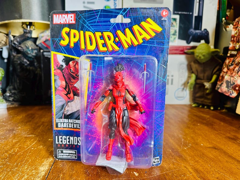 K&ouml;nnte beinhalten: Eine Marvel Spider-Man Legends Series Actionfigur von Elektra Natchios, Daredevil, in einer transparenten Kunststoffverpackung. Die Figur ist rot und schwarz, mit zwei silbernen Sai-Waffen. Die Verpackung zeigt das Spider-Man-Logo und Comic-Kunst.