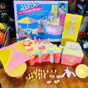 Puede incluir: Un juego de Barbie Ice Cream Shoppe en rosa y amarillo. El set incluye un puesto de helados, accesorios y una caja con el texto "Coolest place in town!" y "Over 50 pieces inside!"