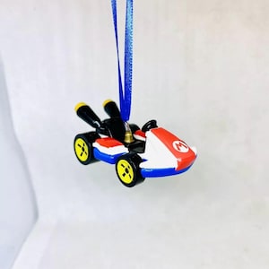 May include: A miniature toy race car ornament with a blue ribbon for hanging. The car features a red and white body with a prominent "M" logo, black accents, and yellow wheels. The overall design is reminiscent of a video game.