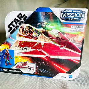 Könnte beinhalten: Eine Star Wars Mission Fleet Delta-7 Jedi Starfighter Spielzeugverpackung. Die Box zeigt einen rot-weißen Sternjäger mit einem grünen Laserstrahl. Das Paket enthält auch eine Anoska Tano Actionfigur. Das Star Wars Logo ist sichtbar.