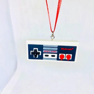 Op de afbeelding: Een witte Nintendo-controller ornament met rode knoppen en een rood lint. De controller heeft de woorden "SELECT" en "START" erop gedrukt. De ornament hangt aan een witte achtergrond.