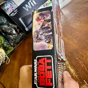 Puede incluir: Una caja de figuras de acci&oacute;n de Star Wars: El libro de Boba Fett. La caja es negra con texto rojo y blanco. El lateral de la caja presenta una imagen de un personaje de la serie. La caja est&aacute; sujeta por una persona.